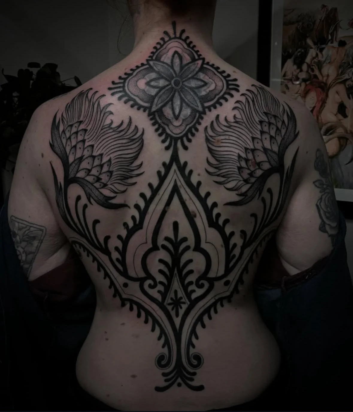 A person's back with tribal-style black tattoos, including a large symmetrical floral pattern on the upper back, two feather-like designs on the shoulders, and a large ornate design extending down the spine.