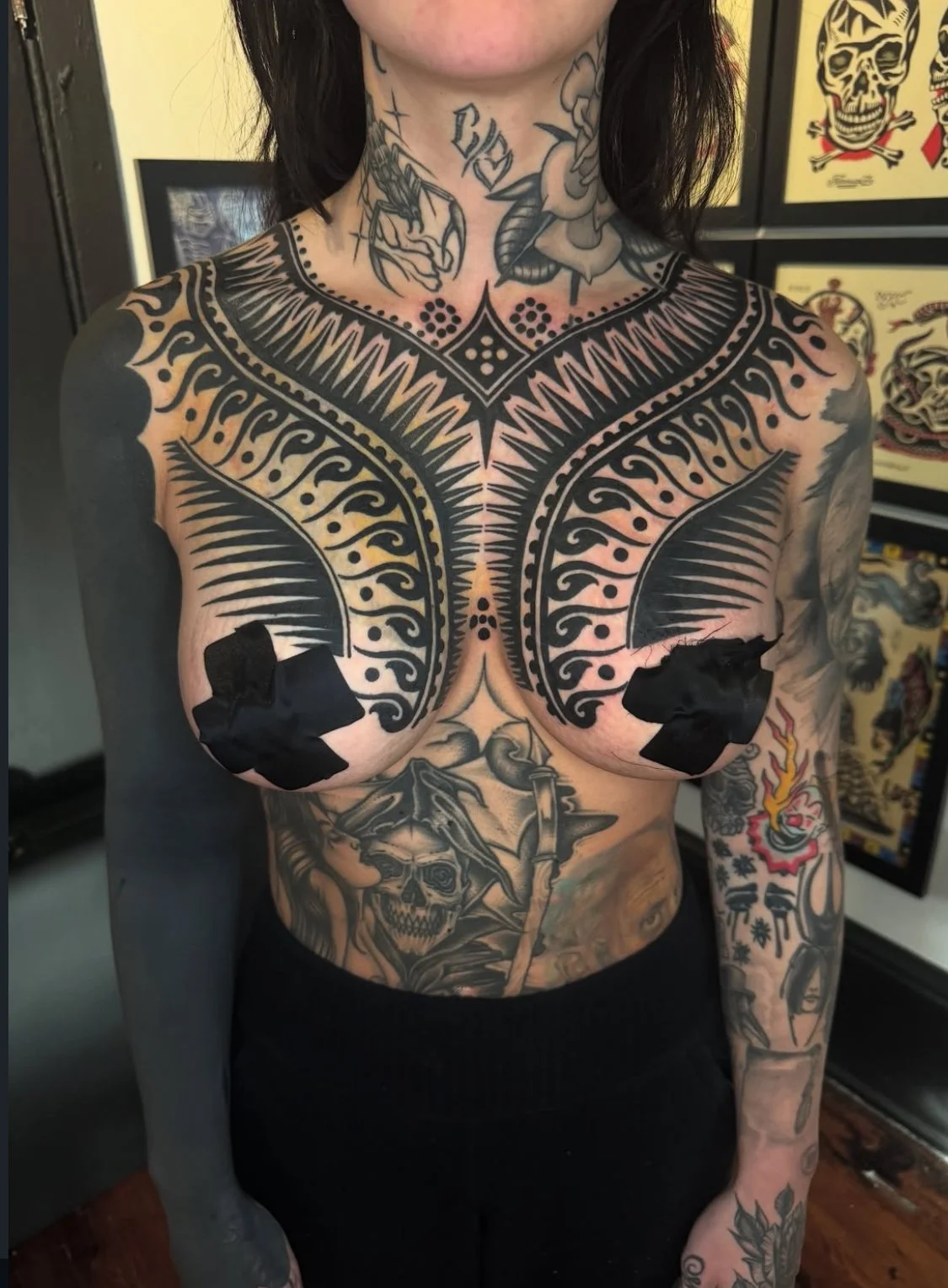 A person with extensive tattoos on their chest, torso, arms, and neck, featuring various black ink designs including geometric patterns, skulls, flames, and floral elements.
