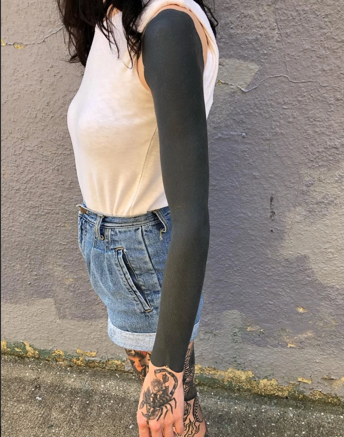 A woman stands against a gray textured wall, wearing a sleeveless beige top, denim shorts with rolled-up cuffs, and has black tattooed sleeves on her arms and a scorpion tattoo on her hand.