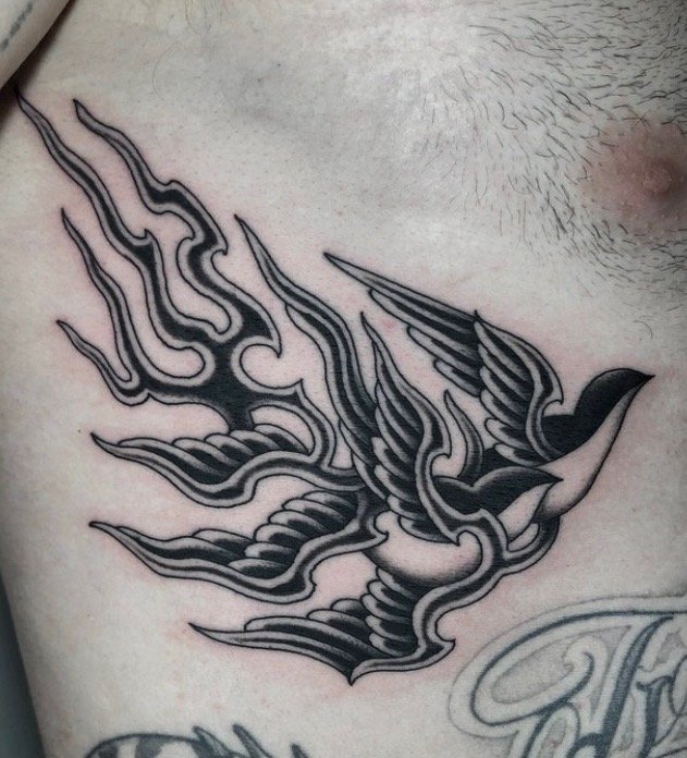 Tattoo of a stylized bird with intricate black and gray lines, located on the person's chest near the shoulder.