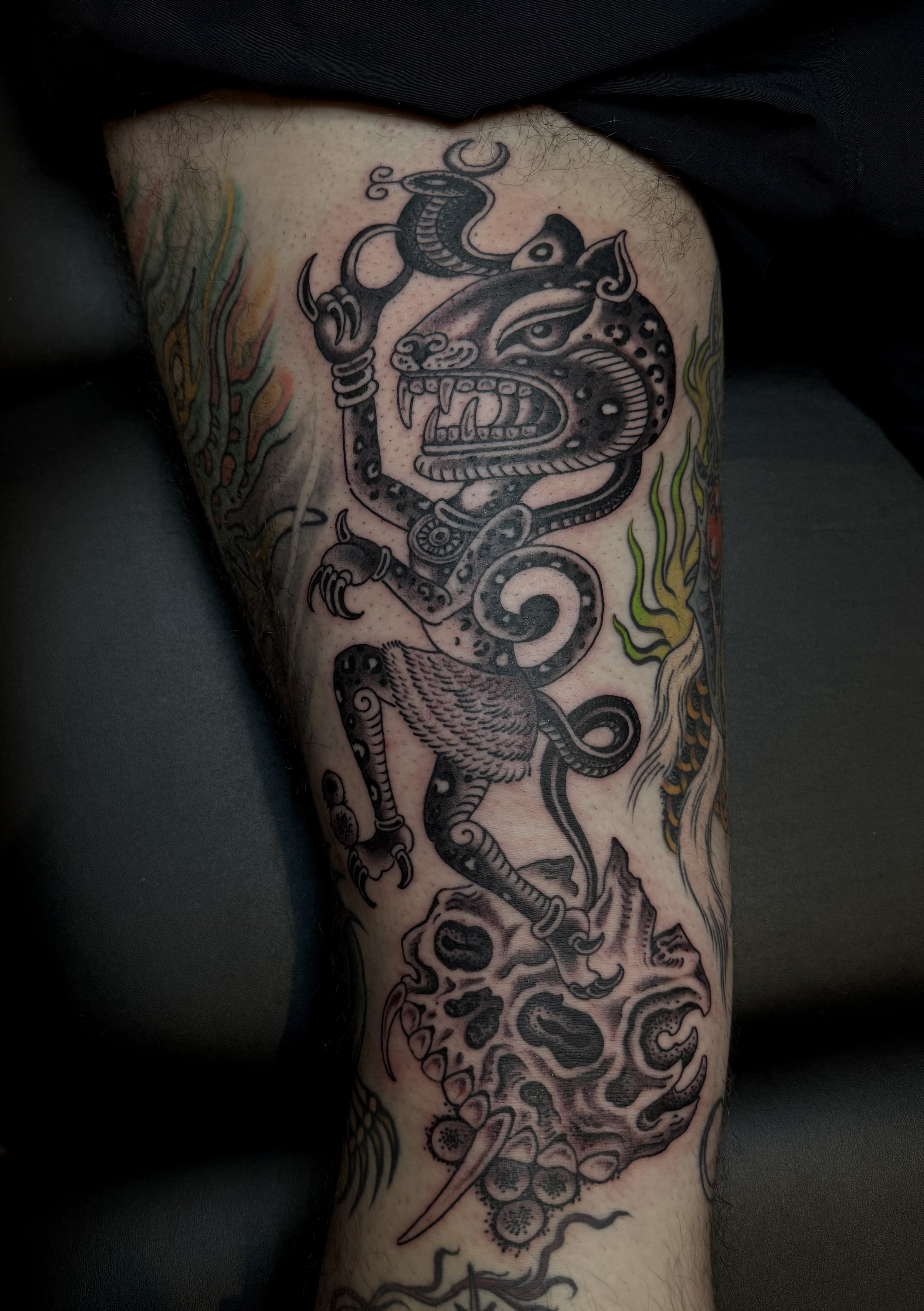 Tattoo of a fierce lion with a snake wrapped around its head, holding a sword in one paw and standing on a rock with flames and abstract designs surrounding it.