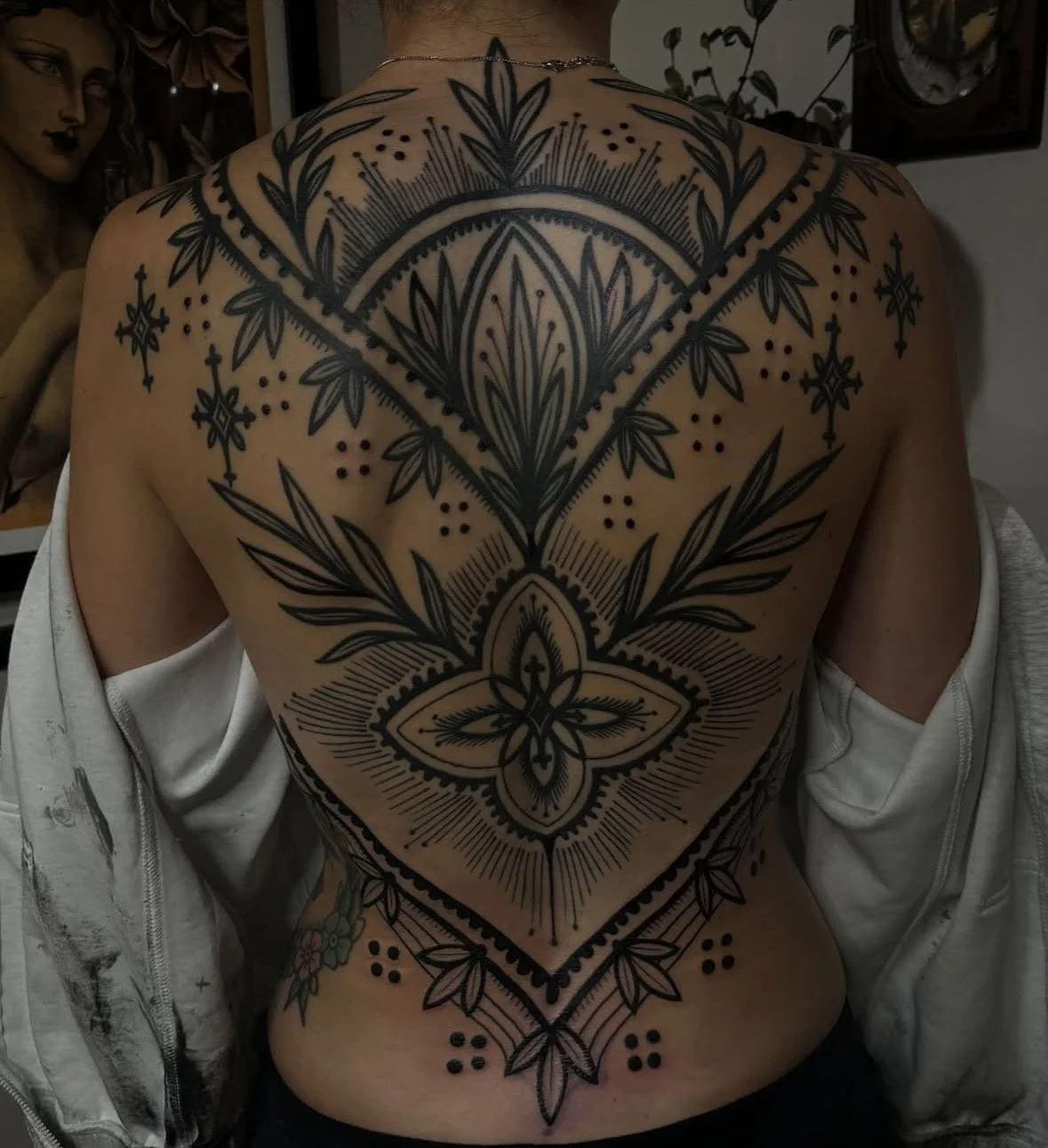 A person's back with a large black geometric and floral tattoo covering the shoulders, upper back, and part of the lower back.