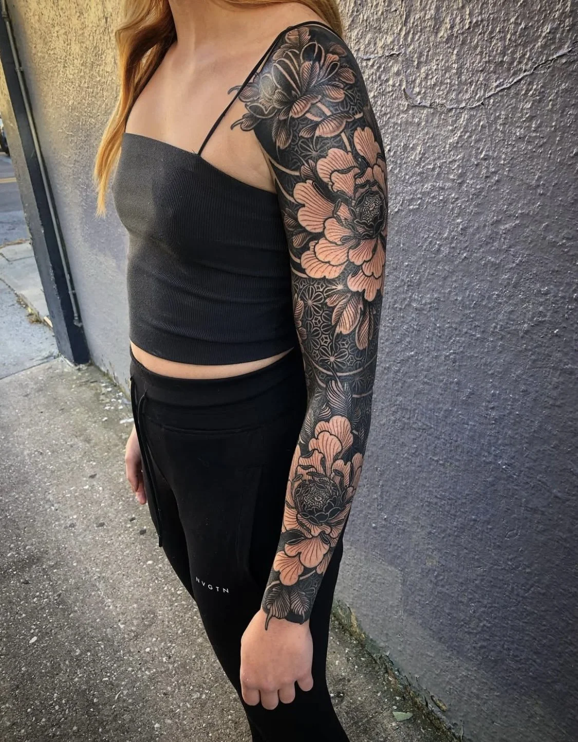 A woman with a large floral tattoo on her left arm, wearing a black tank top and black pants, standing outdoors next to a textured gray wall.