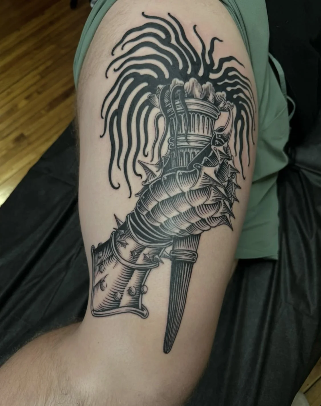 Tattoo of a medieval helmet with a dagger pierced through it, surrounded by exaggerated black lines resembling hair or flames, on a person's upper arm.