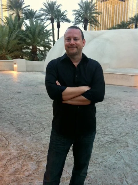 Man standing outdoors with crossed arms, palm trees, and a large white sculpture in the background, in an urban setting with tall buildings.
