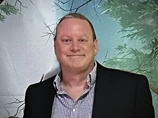 A man with short hair and a friendly smile wearing a checkered shirt and a dark blazer standing in front of a nature-themed backdrop with trees and greenery.