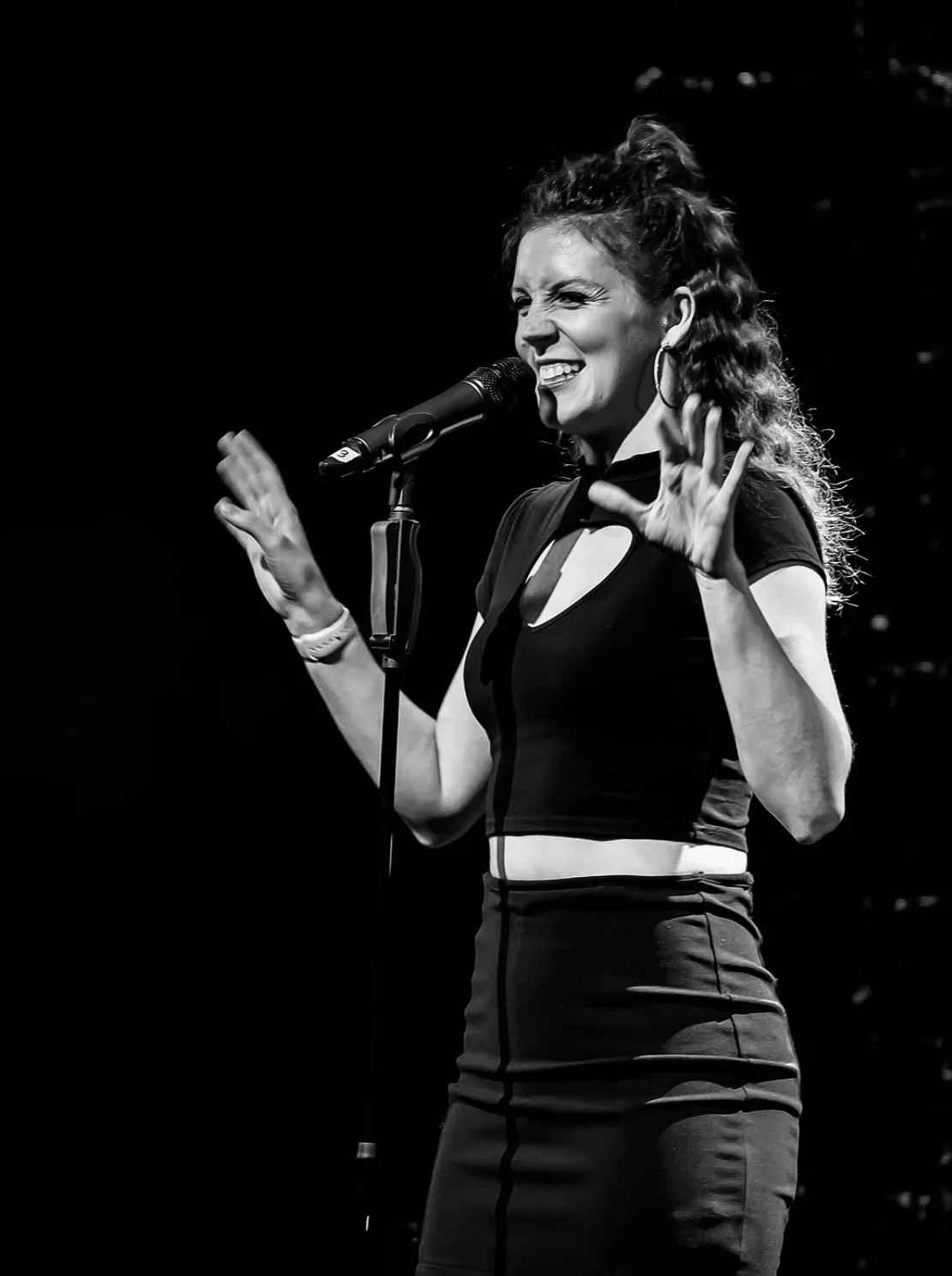 Schwarz-Weiß-Foto einer jungen Frau beim Singen auf einer Bühne, die den Kopf gegen den Mikrofon hält und eine Hand in die Luft hebt.