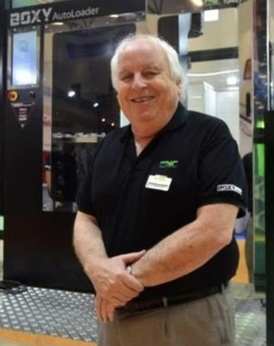 A smiling man with white hair standing in front of a vending machine at an electronics store, wearing a black shirt with store logos.