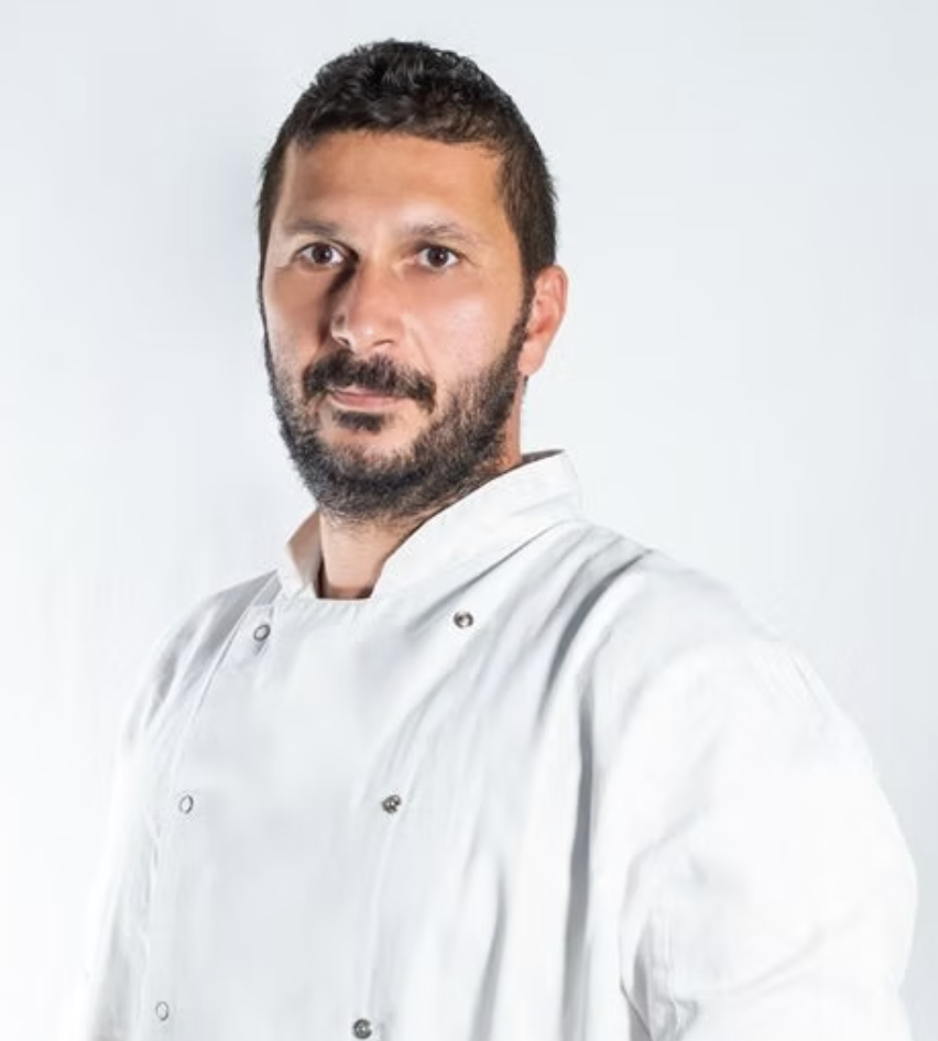A man with dark hair and a beard wearing a white chef's coat against a plain light gray background.