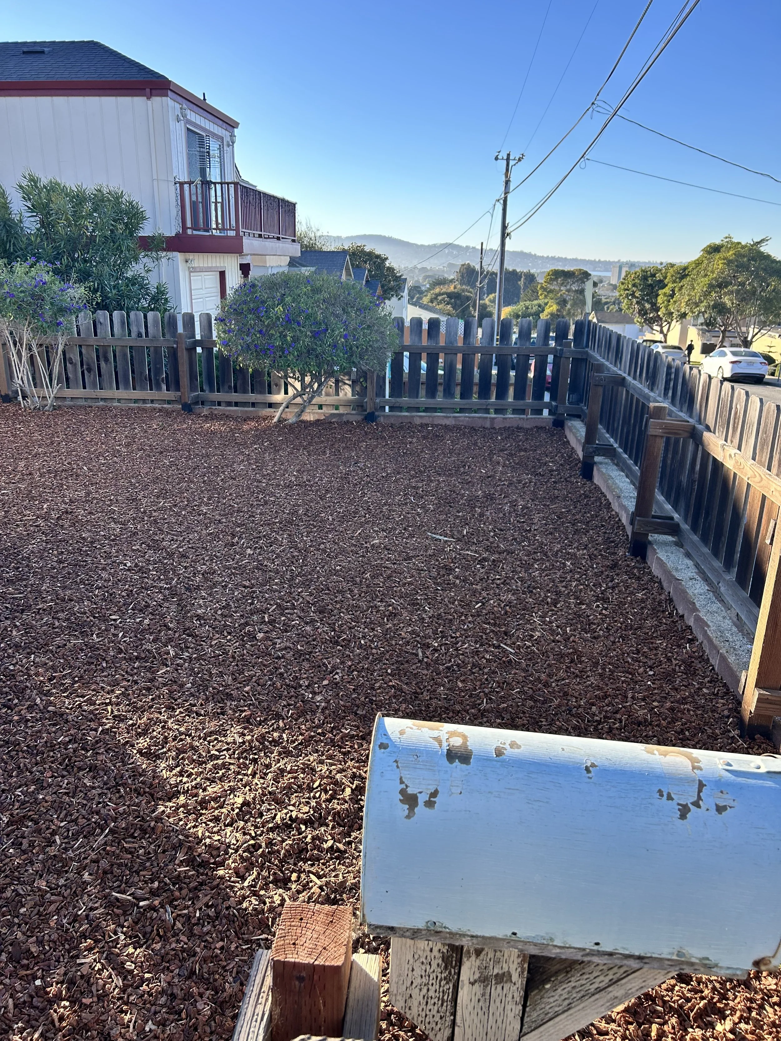 Backyard with wood-chip ground, fenced perimeter, small bush, neighboring houses, parked cars, power lines, and a metallic object in the foreground.