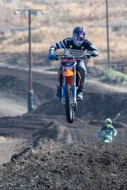 An off-road motocross rider in mid-air jumps over dirt track with another rider in background and power lines in the distance.