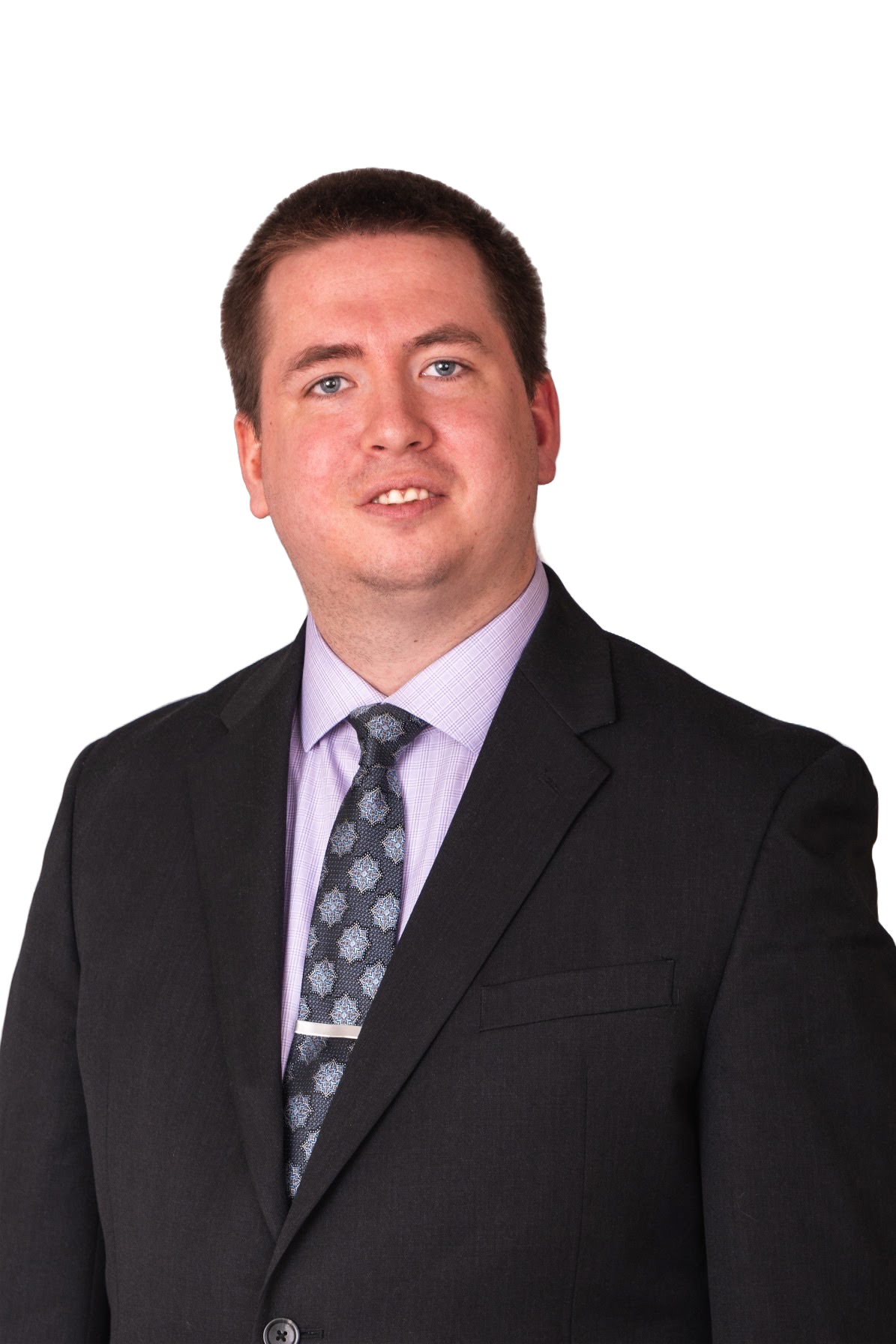 A man with short brown hair, blue eyes, and light skin wearing a black suit, a light purple shirt, and a patterned tie, standing against a dark background.