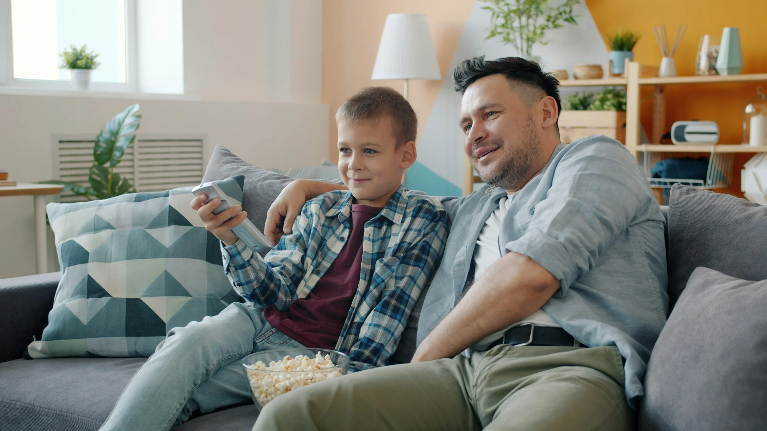 A man and a boy sitting on a couch watching TV, with the boy holding a remote control and a bowl of popcorn in front of them.