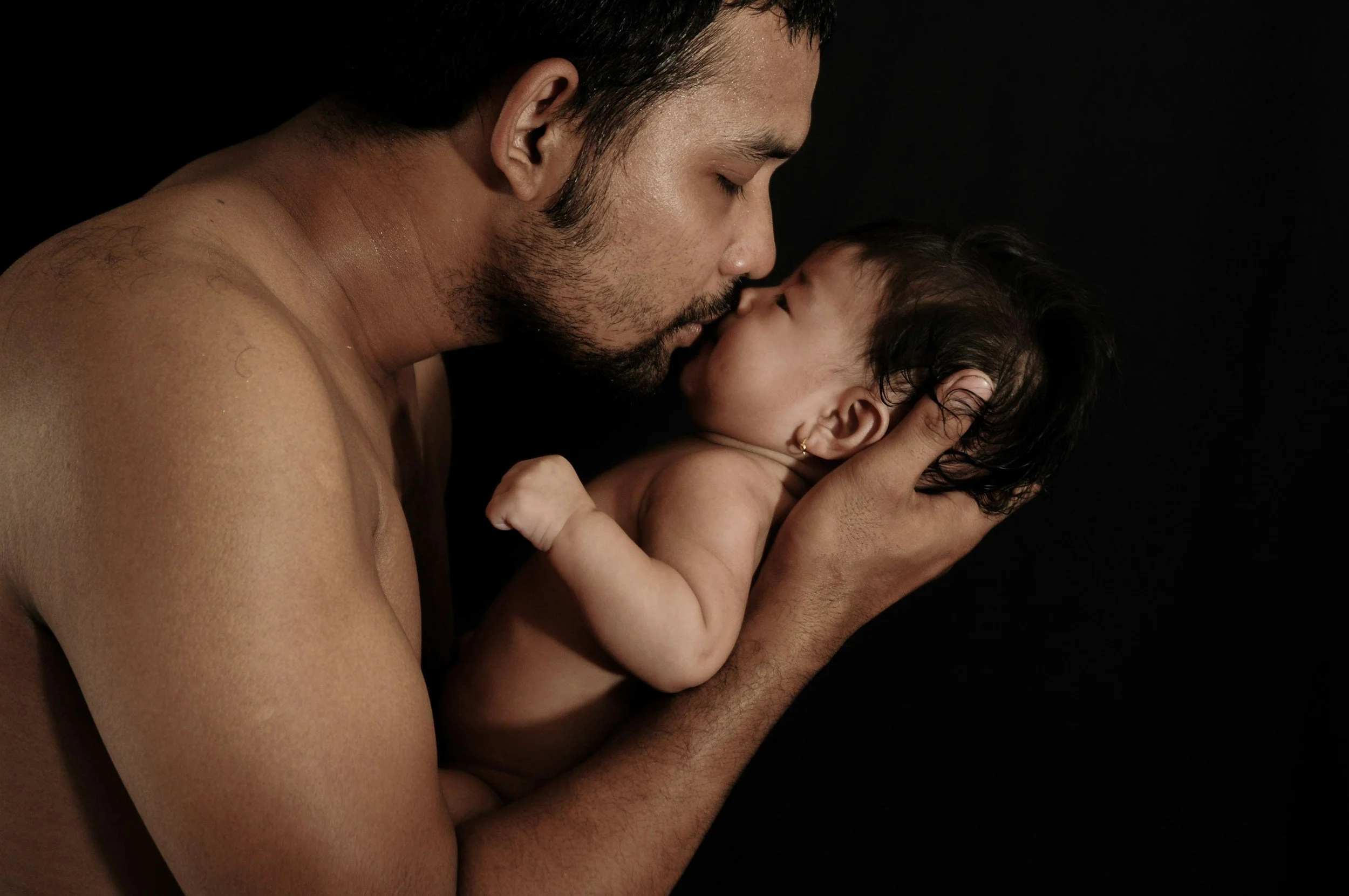 A shirtless man with a beard holding a sleeping baby in his hands, touching foreheads against a black background.