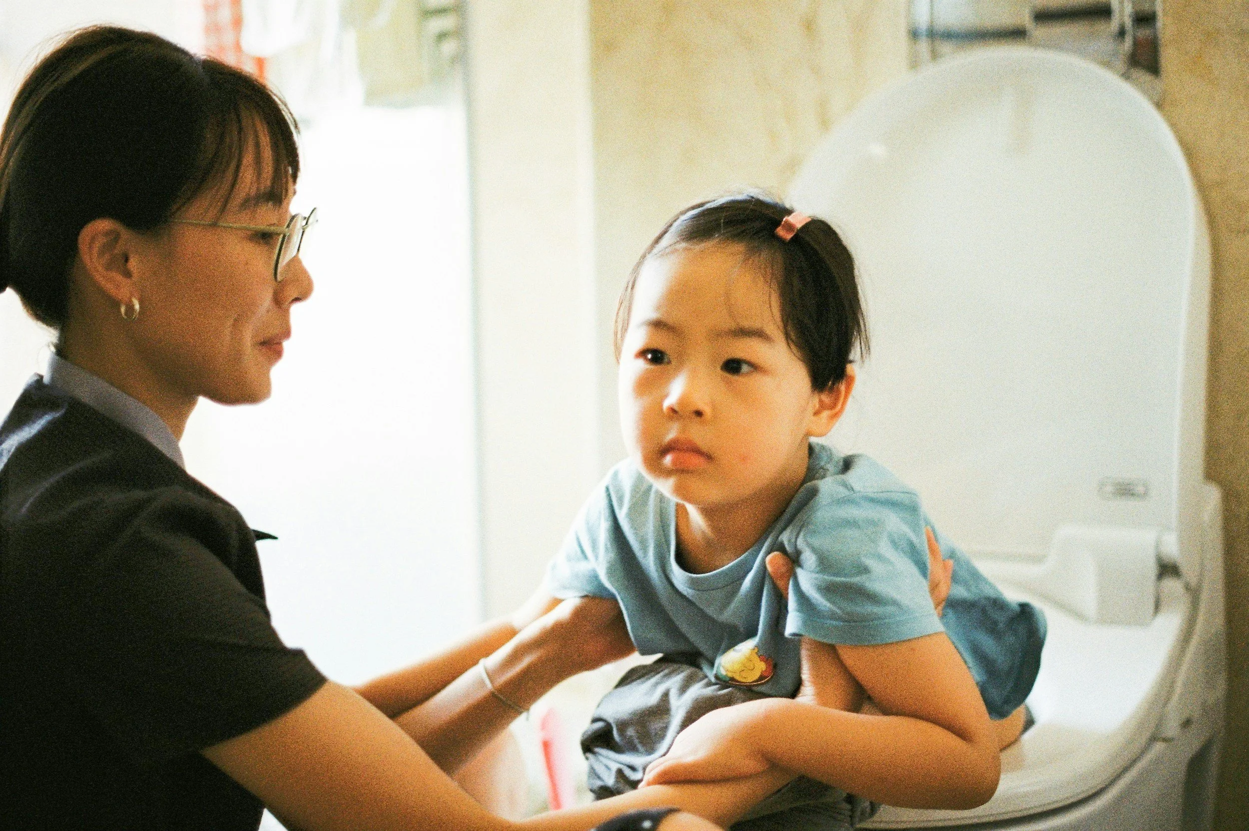 A young girl with a concerned expression sitting on a toilet, being comforted by a woman who appears to be a healthcare worker or nurse.