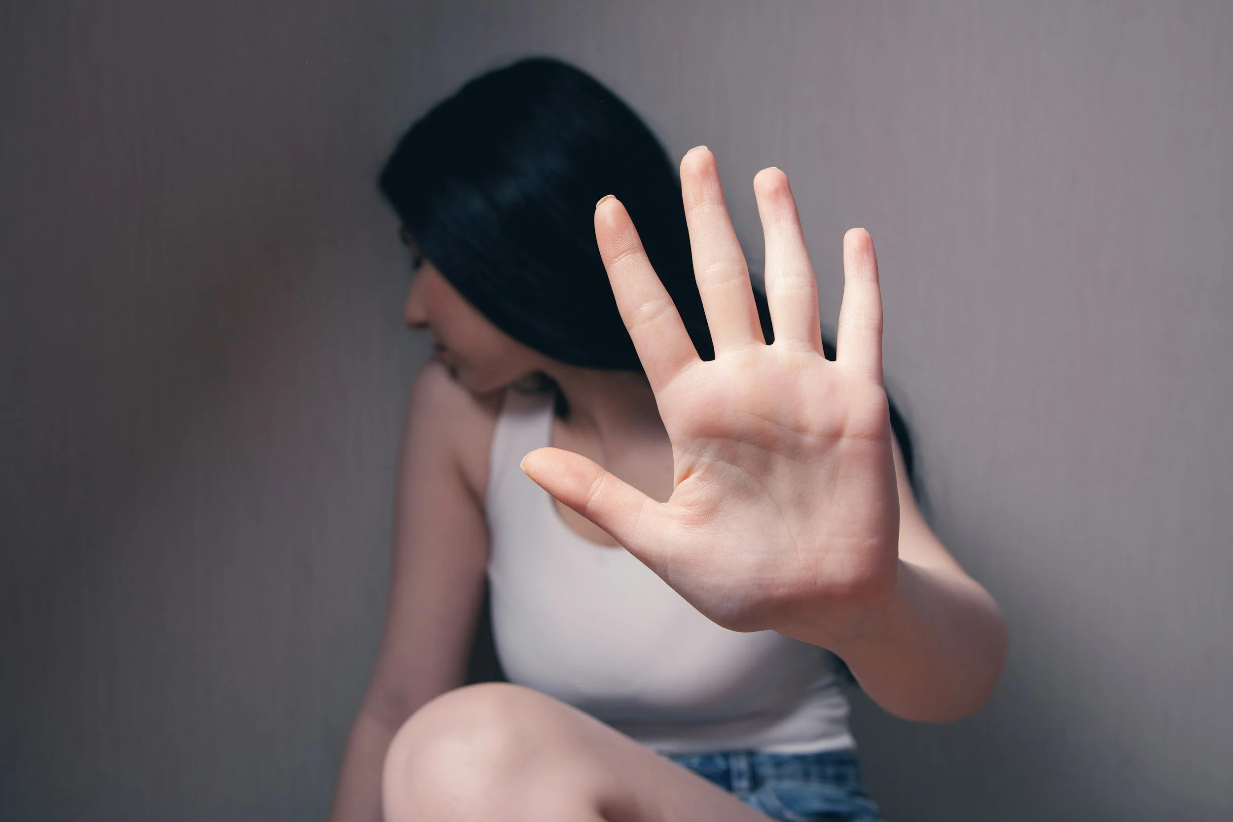 A woman with black hair reaching out her hand toward the camera, facing sideways, with her face turned away, wearing a sleeveless white top and sitting against a plain wall.