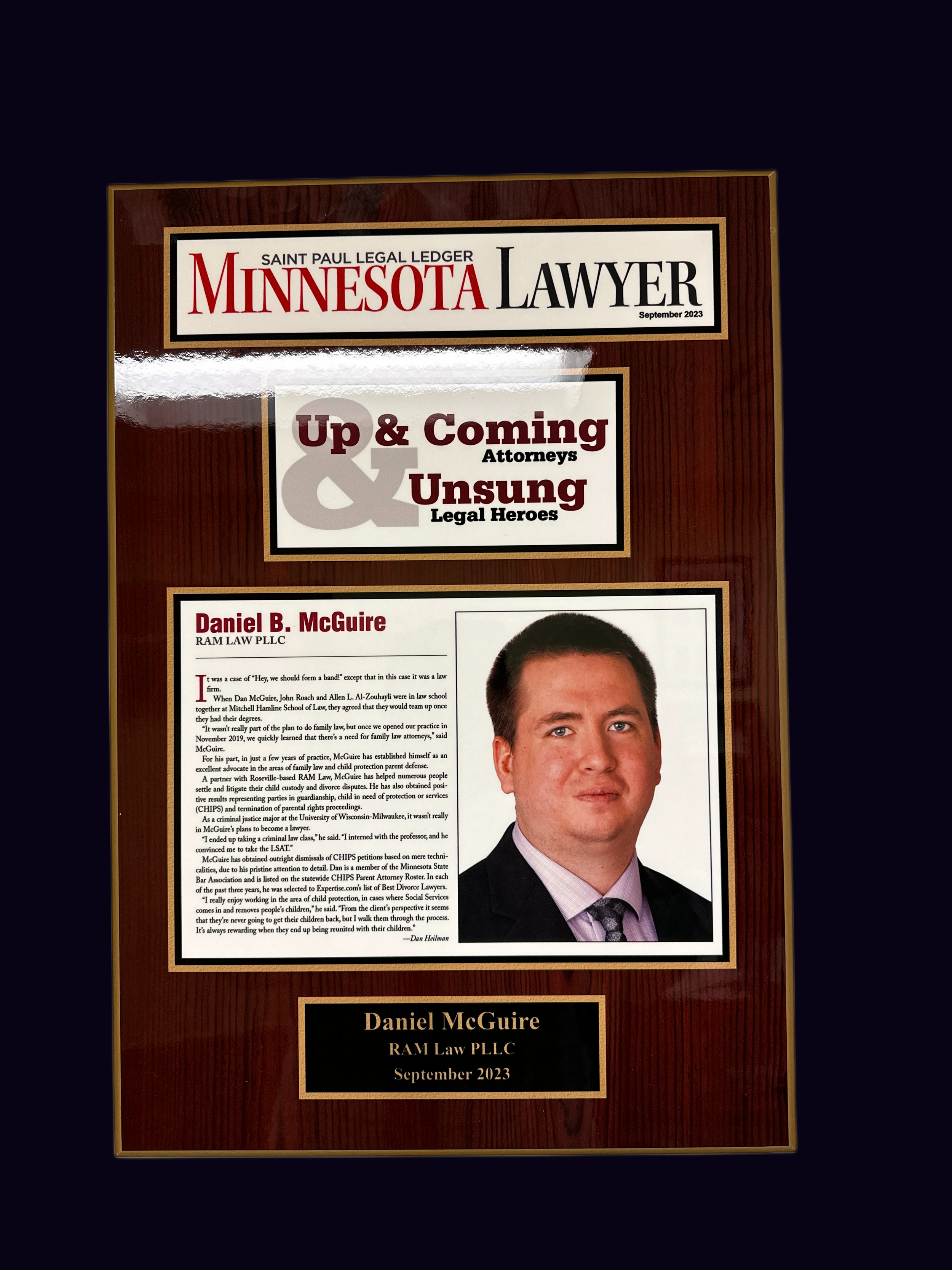 A wooden plaque with a magazine cover design featuring Daniel B. McGuire from RAM Law PLLC, September 2023, with a portrait photo of him in a suit and tie, and text describing his legal background.