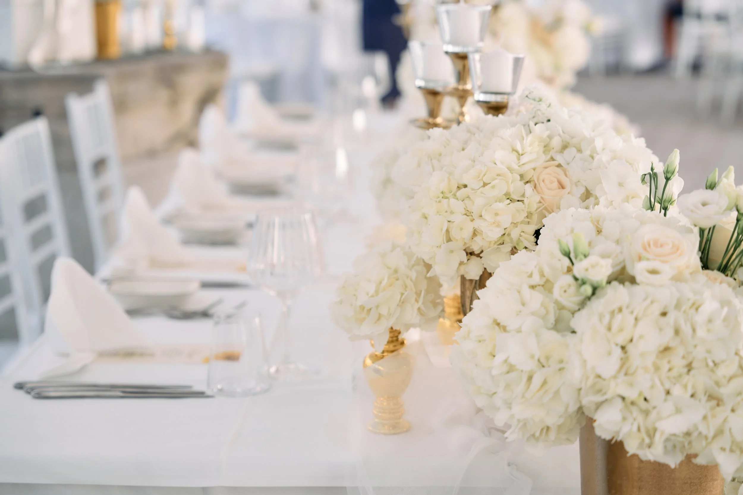 Elegant banquet table decorated with white and cream floral arrangements, gold candelabras, wine glasses, and white napkins, set for a formal event or wedding reception.