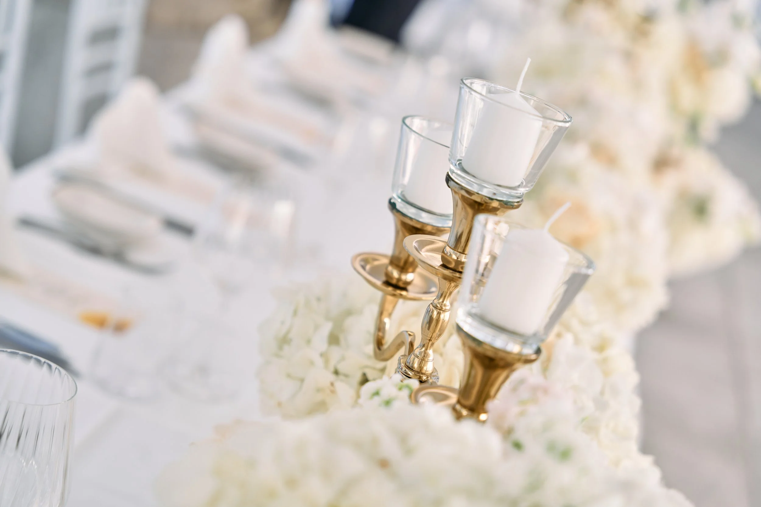 Multiple white candles in glass holders on a flower-decorated table at a wedding or special event.