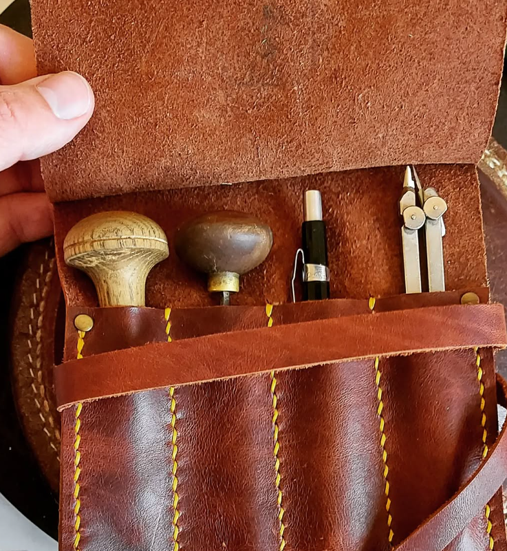 A leather tool roll containing three tools: a wooden-handled mushroom-shaped tool, a darker mushroom-shaped tool, and a metal compass, with a pen also inside the roll.