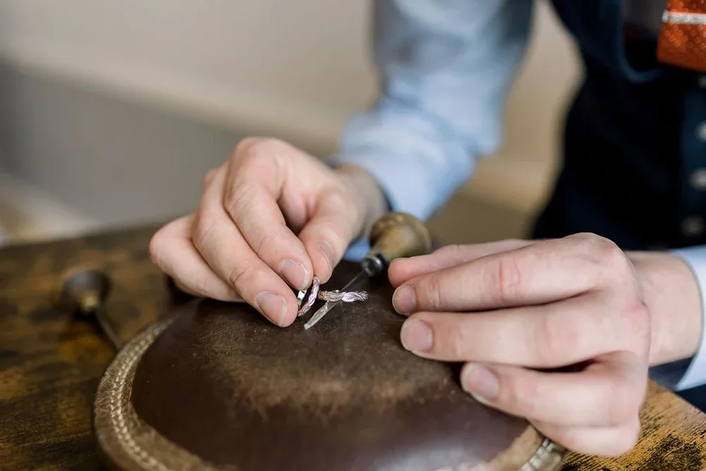 Close-up of a person using a small metal tool to craft or engrave on a piece of leather with a wooden-handled stylus, on a wooden work surface.