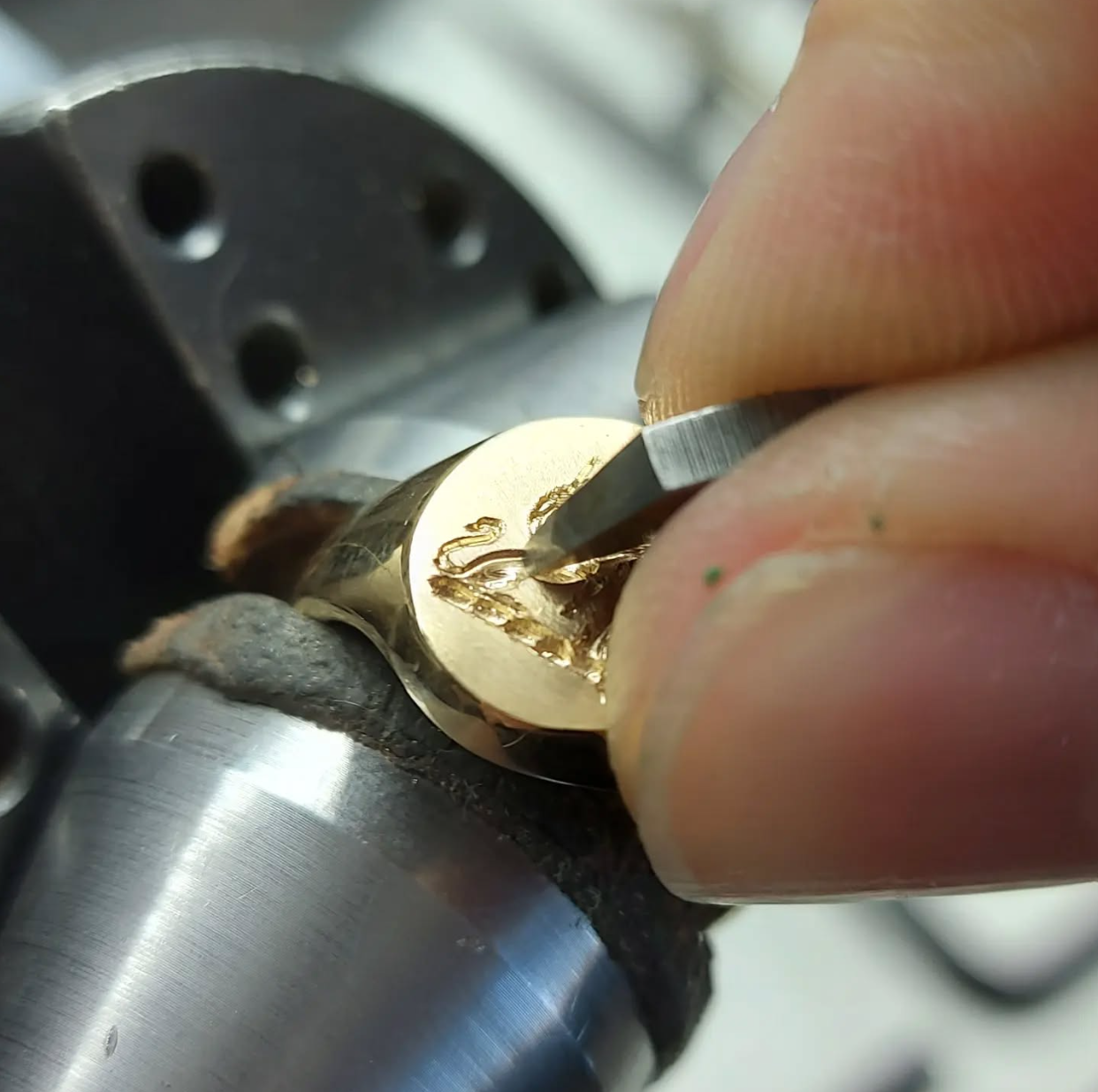 Close-up of a person engraving a name or design onto a gold ring using a small engraving tool, with a watch or machinery in the background.
