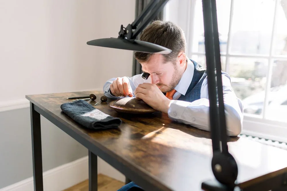 A man working on a watch at a wooden workbench in a well-lit room, with tools and a towel nearby.