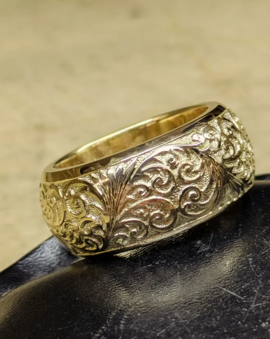 A gold ring with intricate floral and scroll patterns, featuring embedded small diamonds, resting on a black surface.
