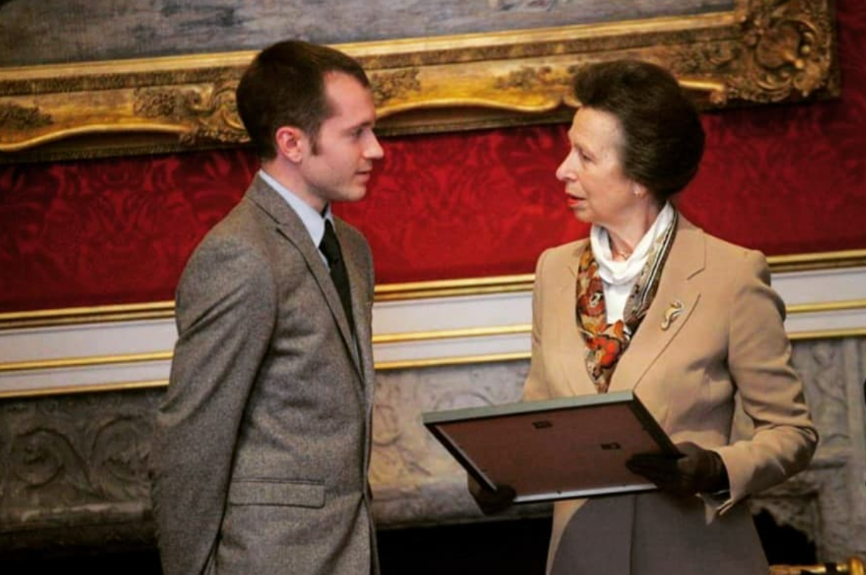 A young man in a gray suit and an older woman in a beige blazer and patterned scarf engaging in conversation in an elegant setting with ornate decor and red upholstery.