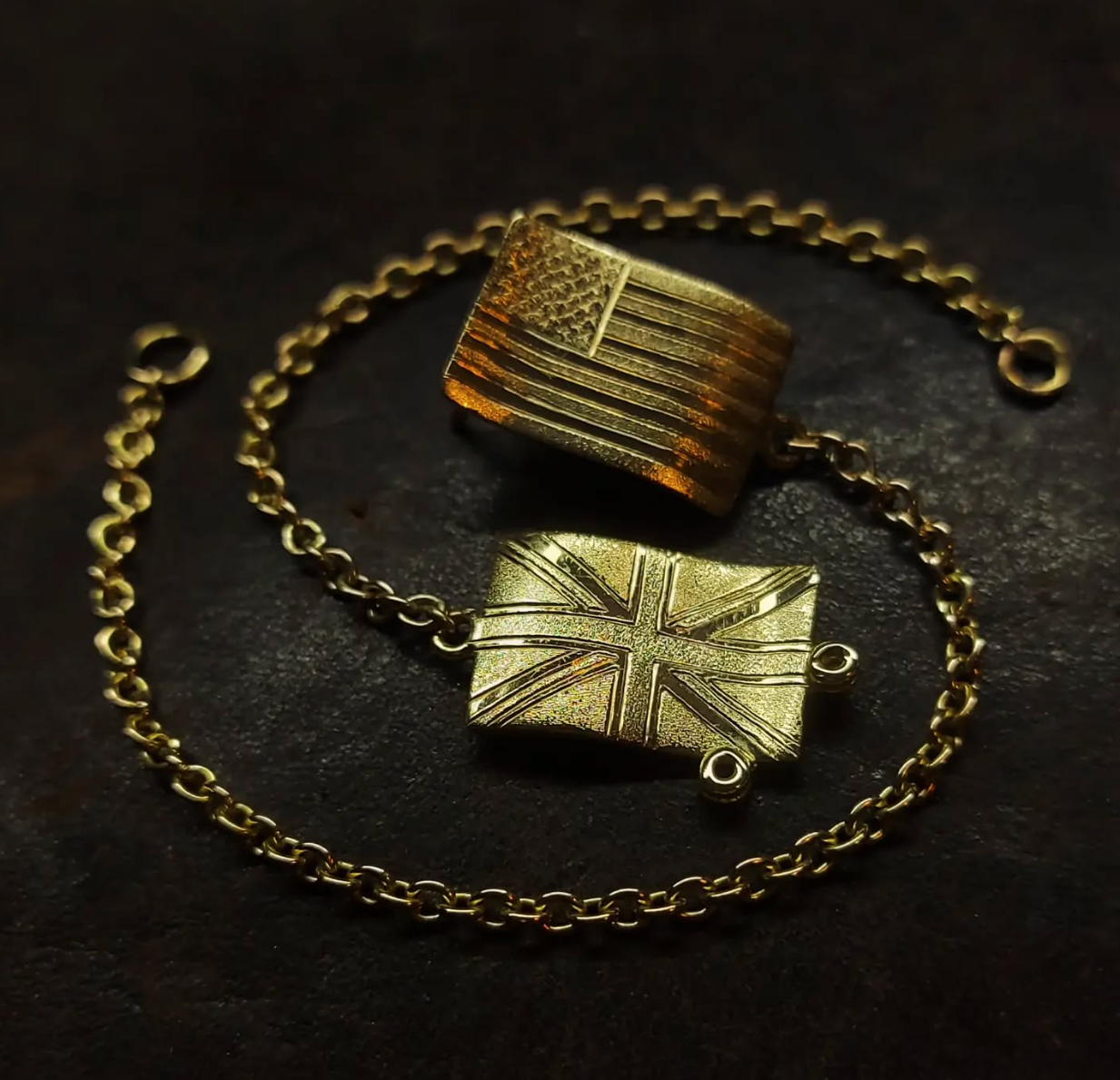 Gold bracelet and two gold charms, one with an American flag design and the other with a Union Jack design, on a dark background.