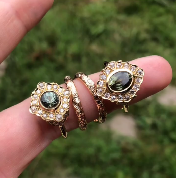 Close-up of three gold rings with crystals and dark central stones, worn on a person's fingers against a green outdoor background.