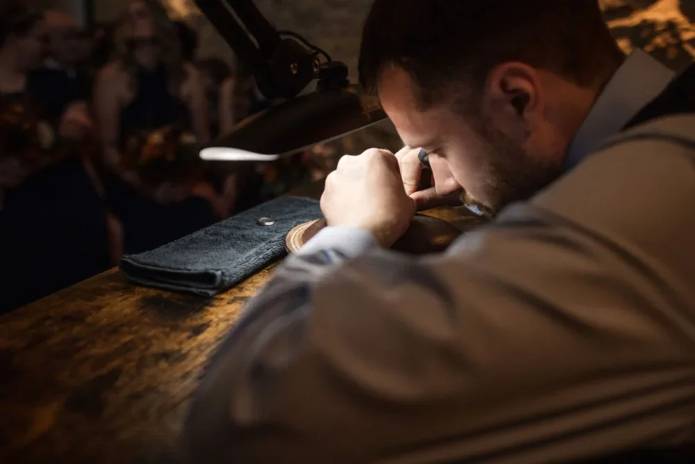 Man sitting at a wooden bar, looking down and inspecting a coin with a magnifying glass under a focused lamp, with a black cloth and a coin holder on the bar