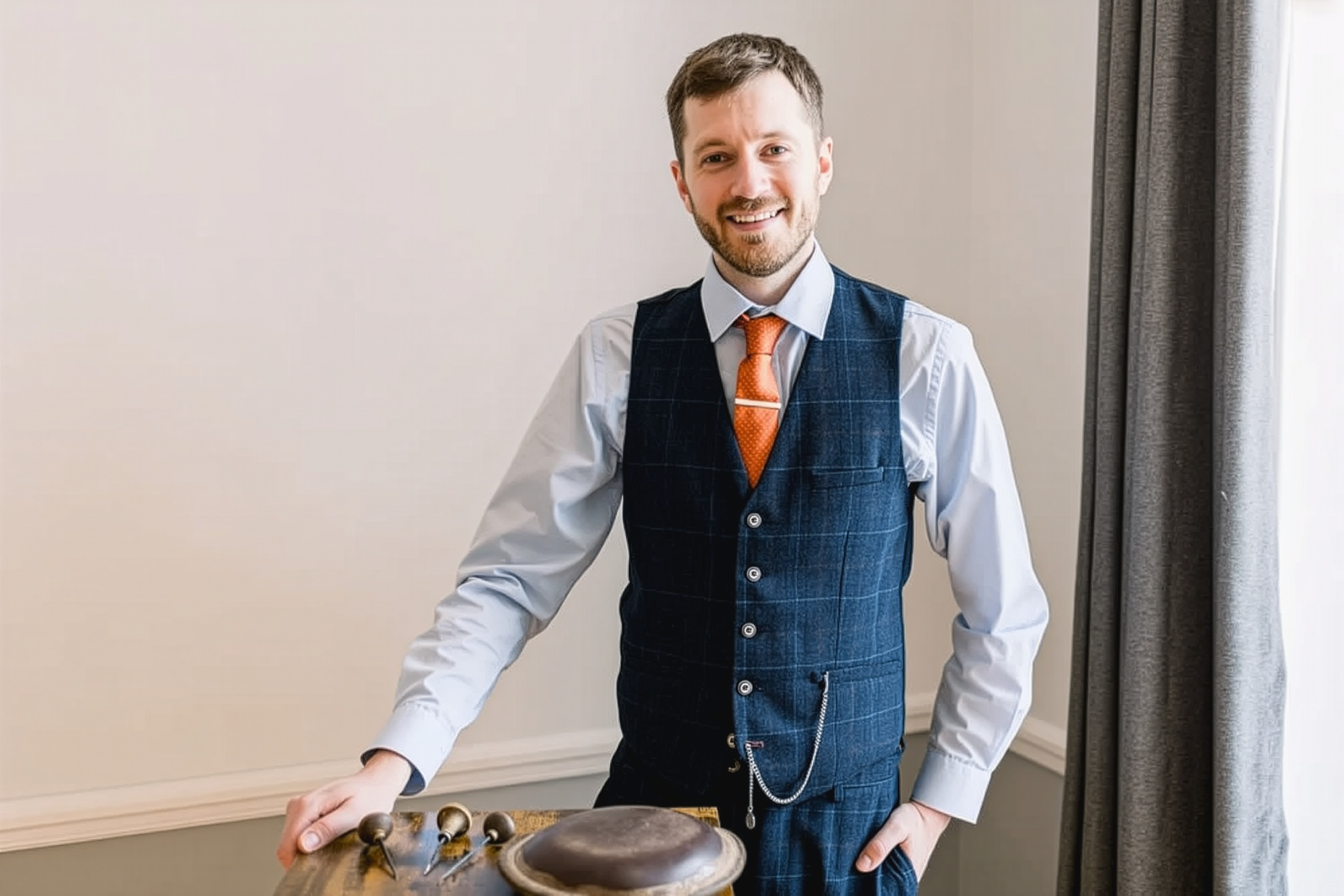 A man in a white shirt, blue waistcoat, and orange tie smiling at the camera, standing near a table with a musical instrument.