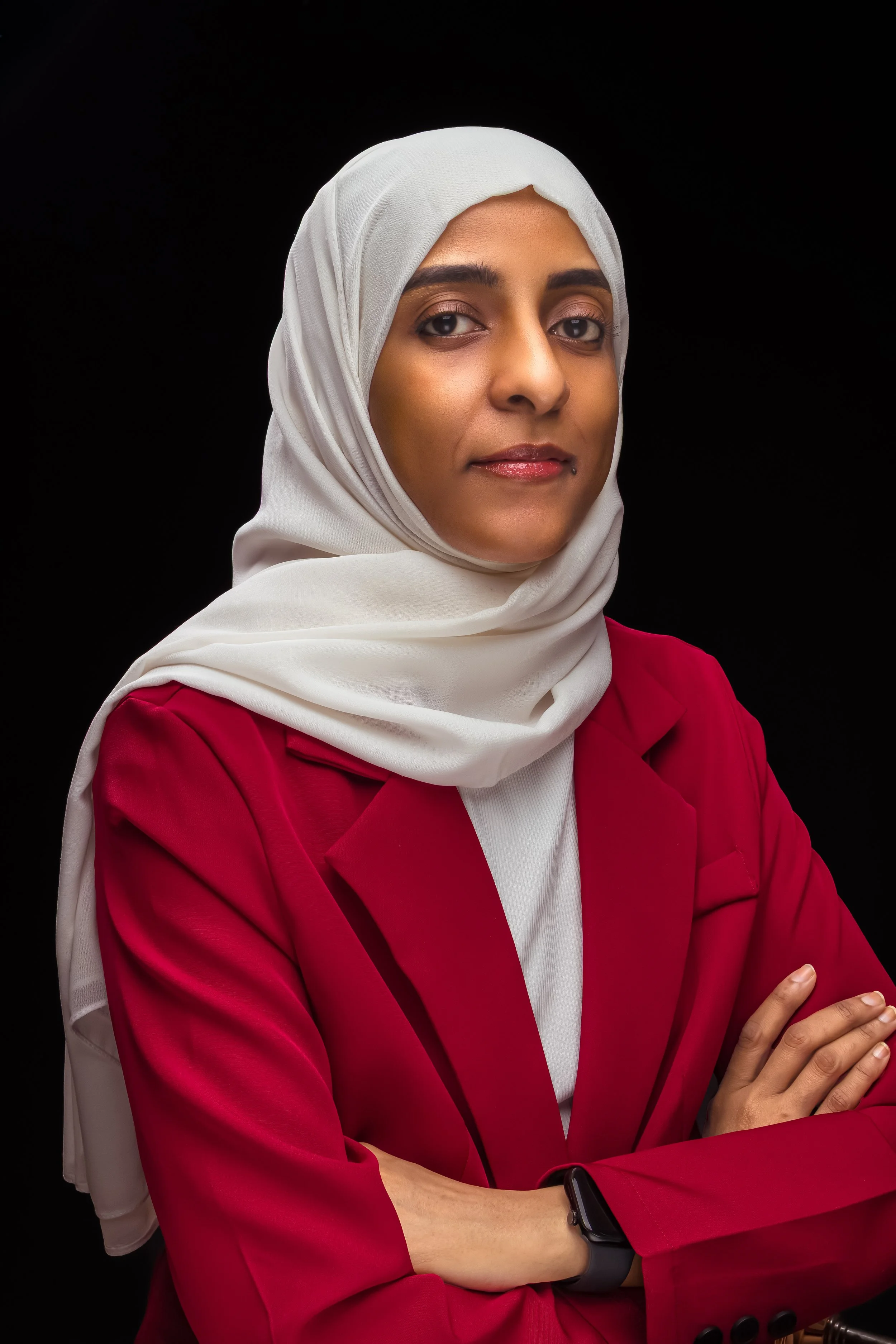 A woman wearing a white hijab and a red blazer, standing with her arms crossed against a black background.