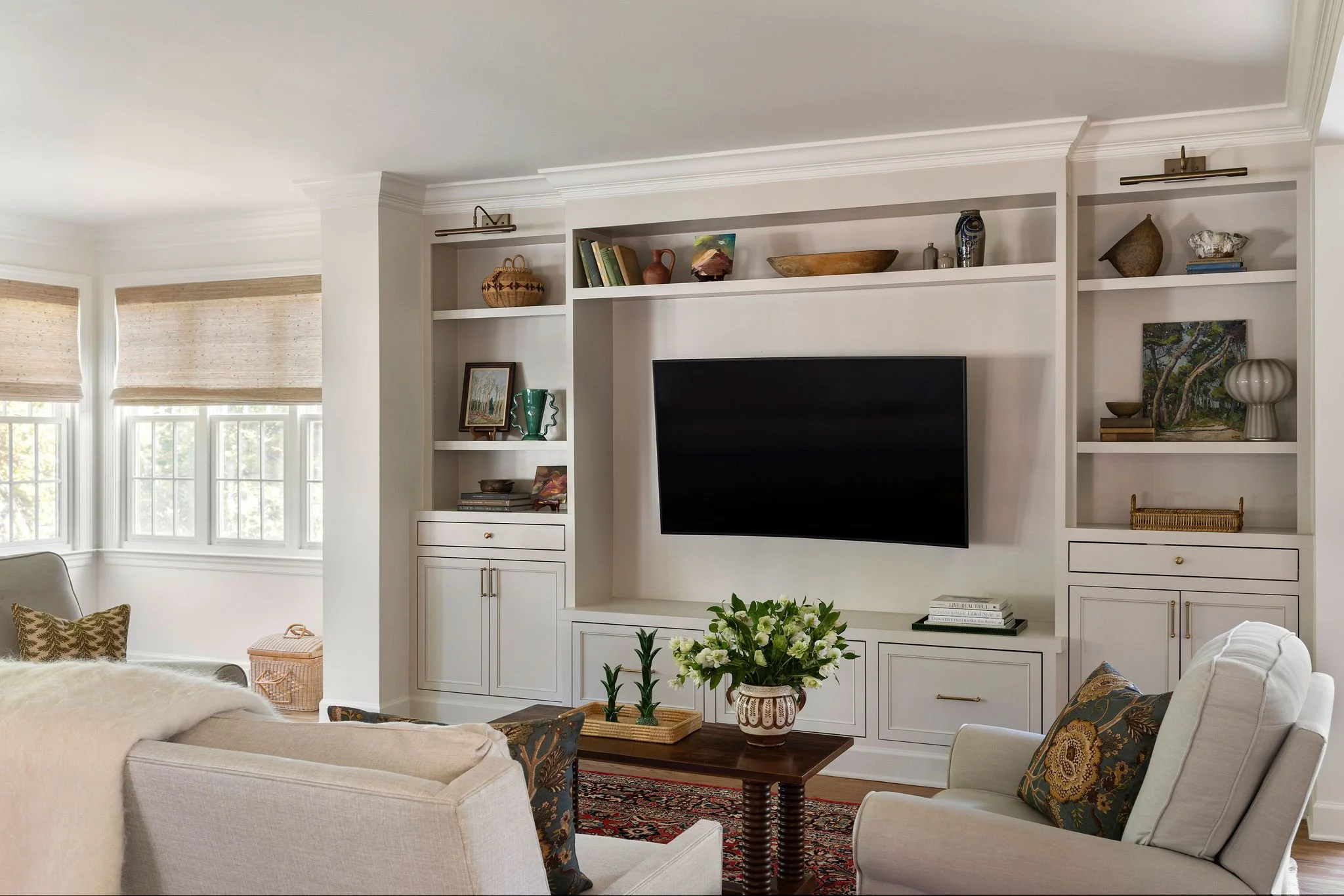 Cream-colored custom living room media wall with integrated shelving, brass picture lights, and a wall-mounted TV above styled cabinetry.
