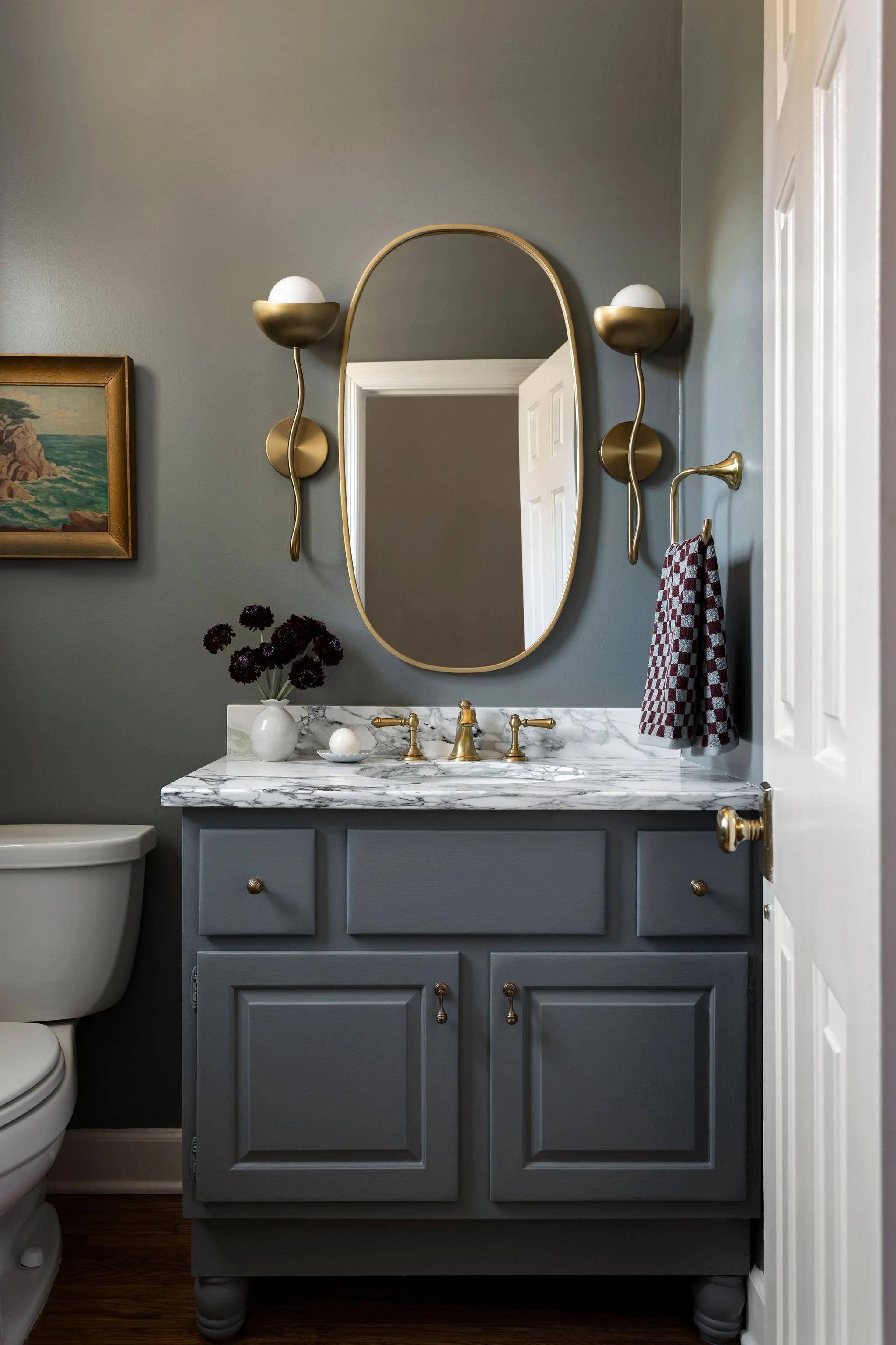 Mood bathroom design with a deep green vanity, marble top, oval gold mirror, and elegant brass wall sconces.