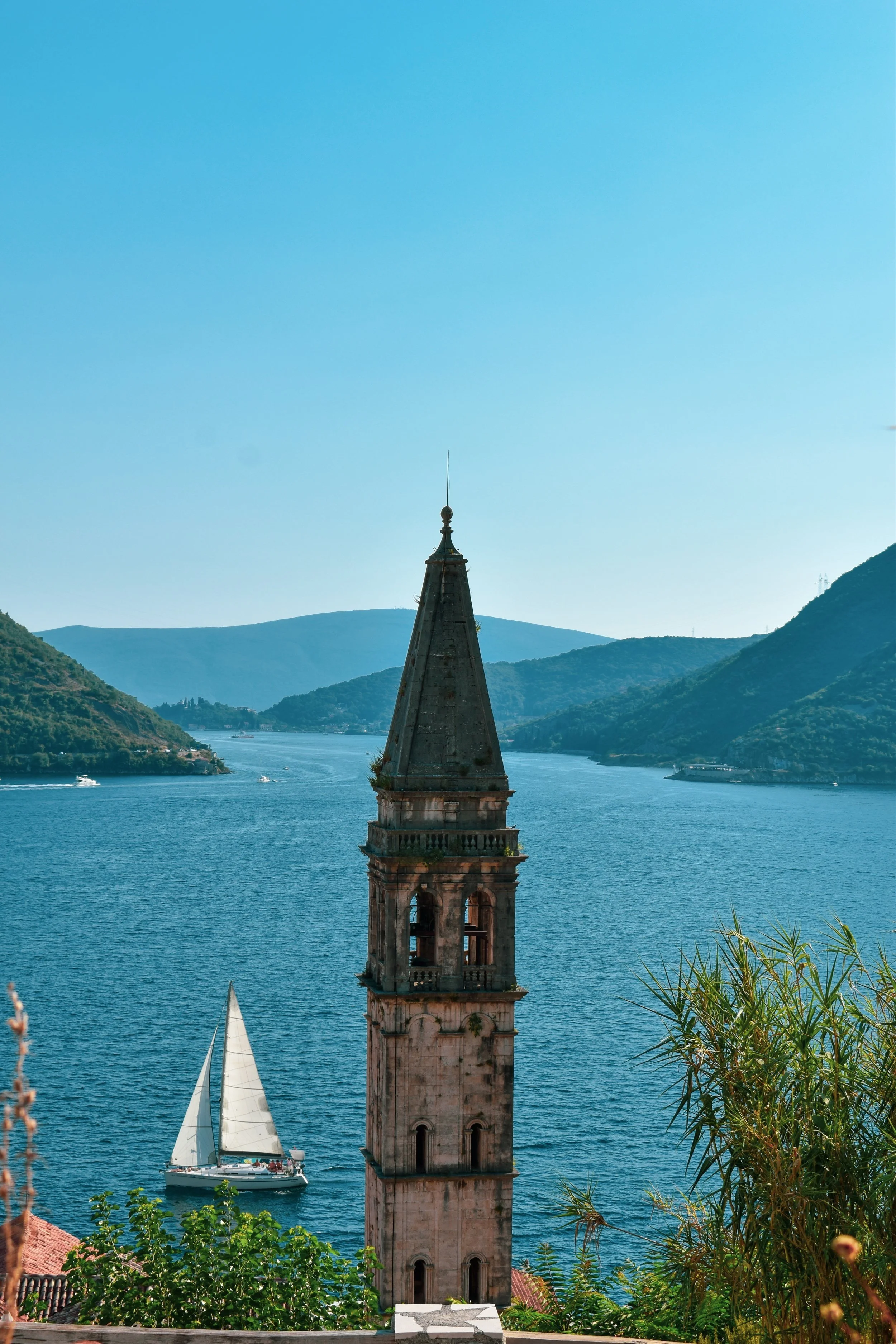 A scenic view of a lake with a sailboat, surrounded by green mountains, and an old church steeple in the foreground.