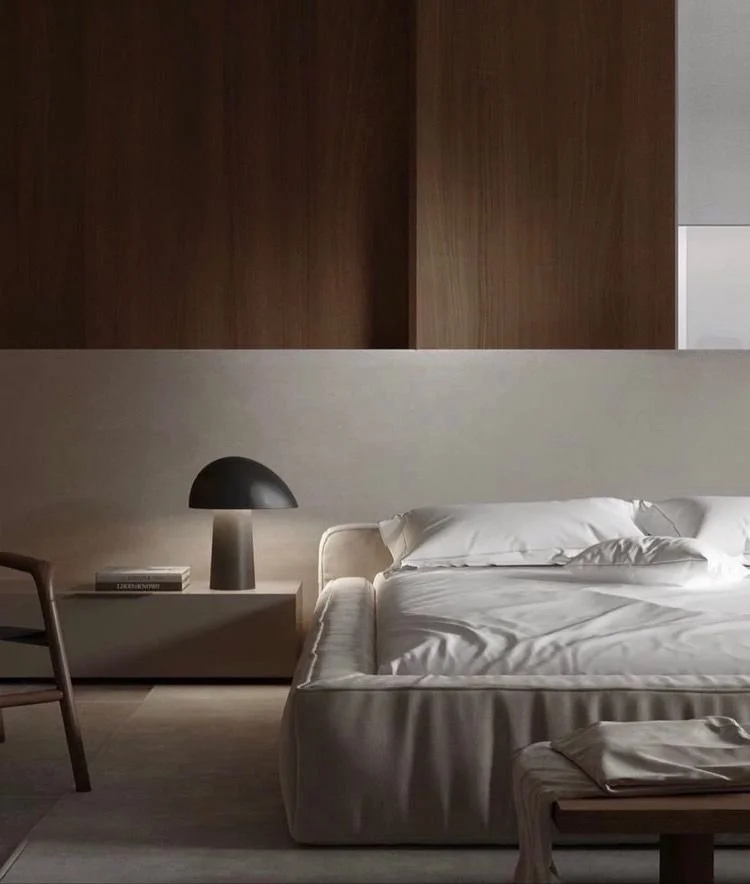 A minimalist bedroom with a white bed, a beige headboard, a bedside table with a black desk lamp and a book, and a wooden chair against a wood-paneled wall.