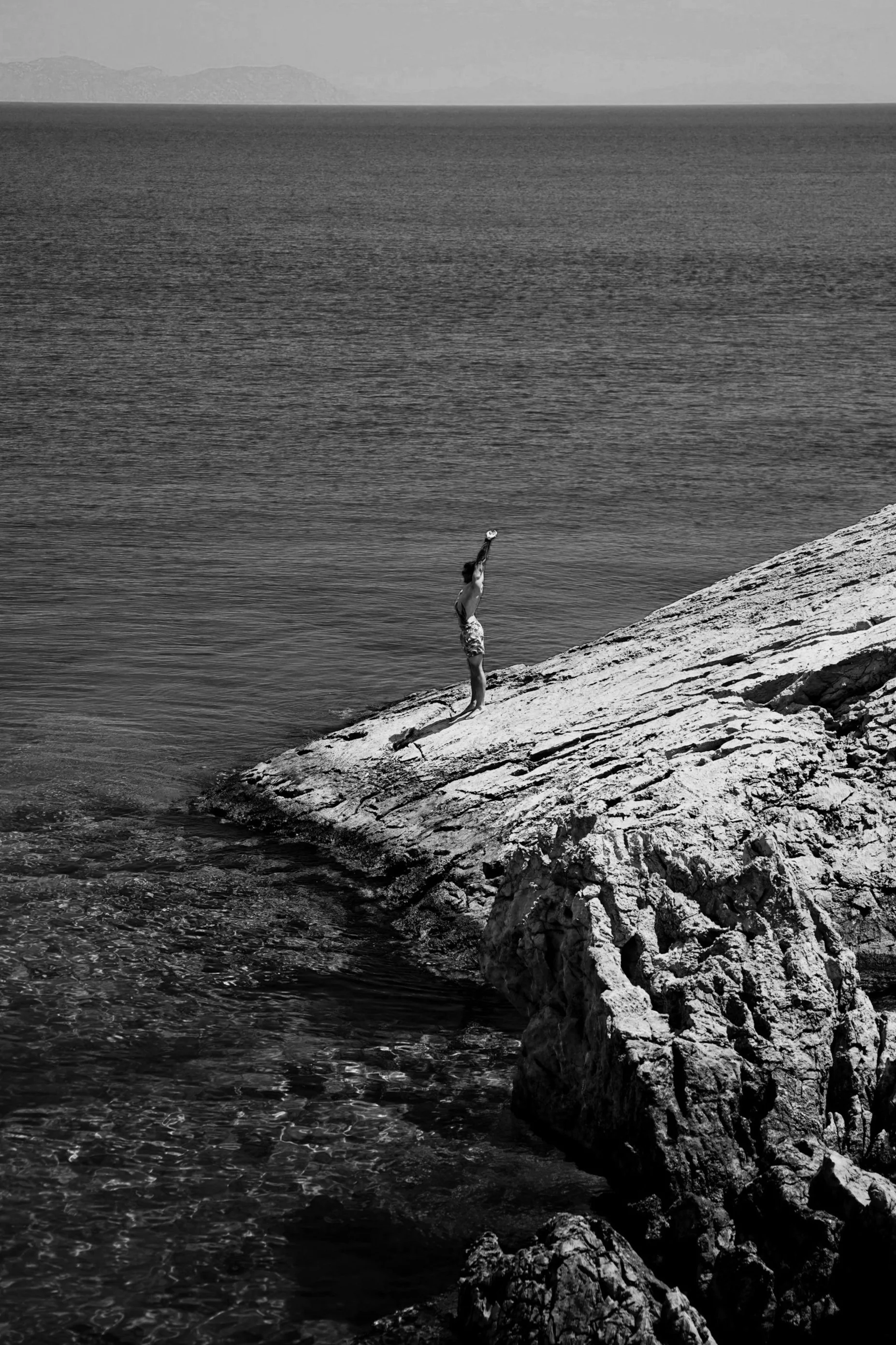 A person standing on a rocky shoreline, performing a handstand with one hand, holding a ball near the water's edge, with an expansive body of water and mountains in the background.