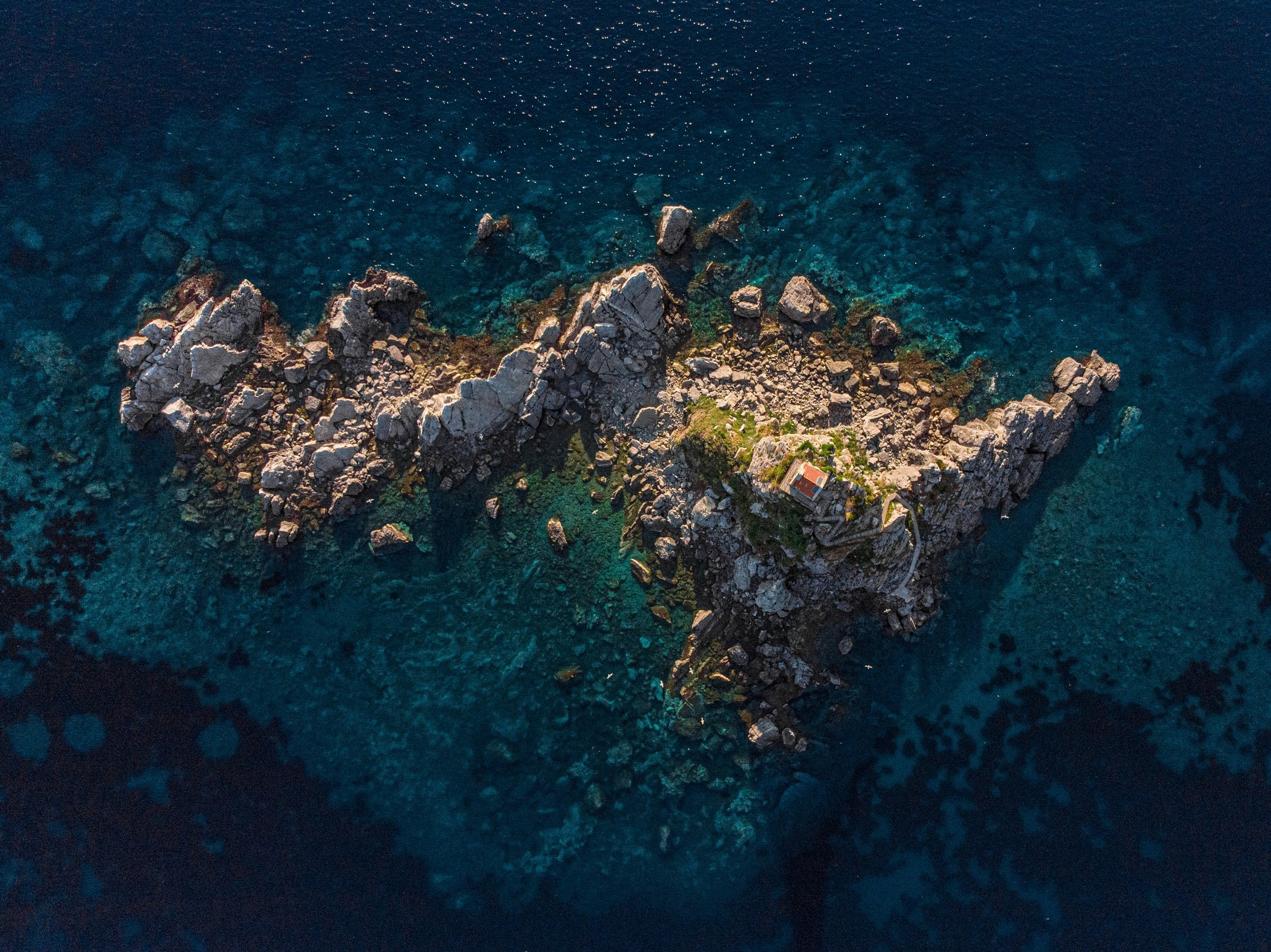 Aerial view of a rocky island surrounded by clear blue ocean water, with a small building and greenery on the island.