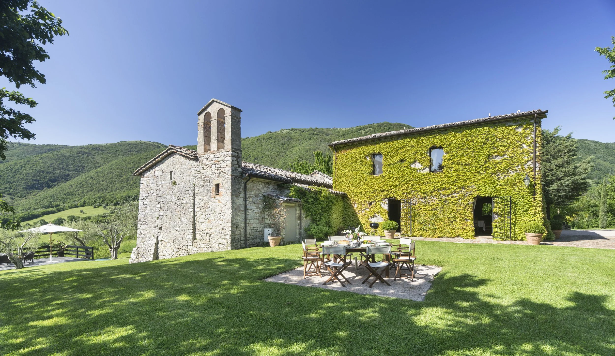A stone and ivy-covered building in a scenic countryside with mountains in the background, outdoor dining table with chairs, a parasol, and green grass.