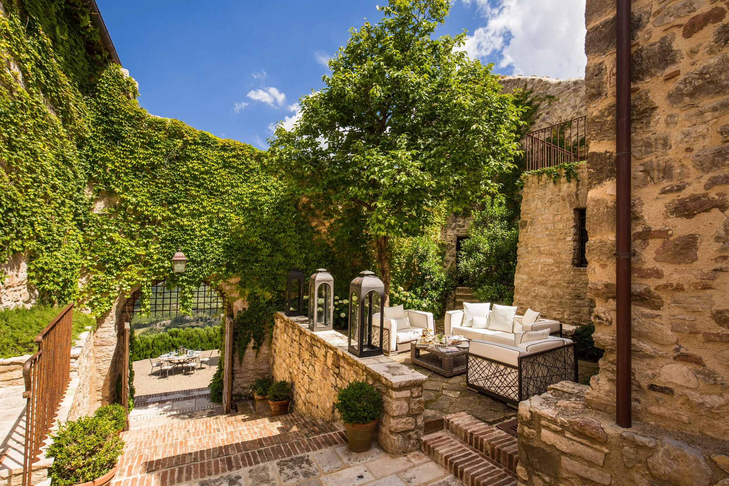 Outdoor patio area with white cushioned seating, potted plants, lanterns, and brick and stone walls, surrounded by green ivy and trees under a blue sky