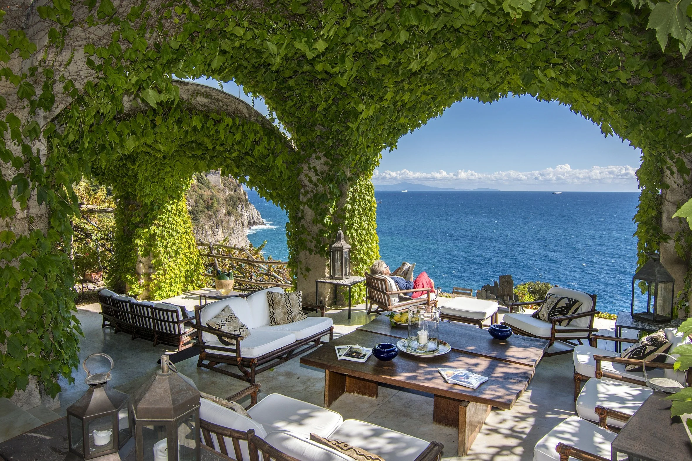 Outdoor patio with white cushioned seating and a wooden table, overlooking the ocean with cliffs and a clear blue sky.