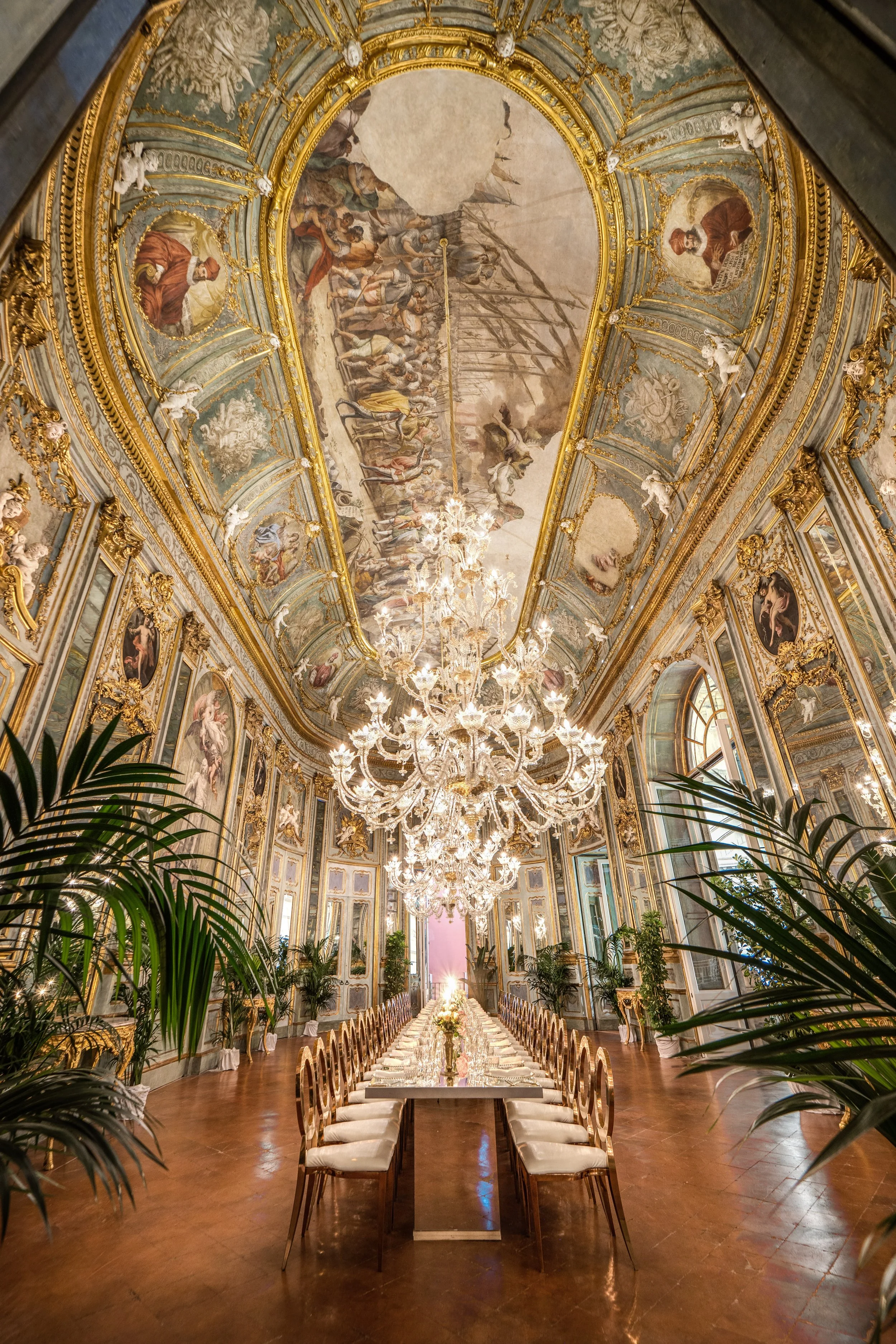 Luxurious, ornate ballroom with high, decorated ceilings, large chandelier, long dining table with chairs, and elaborate gold trim and artwork on walls.