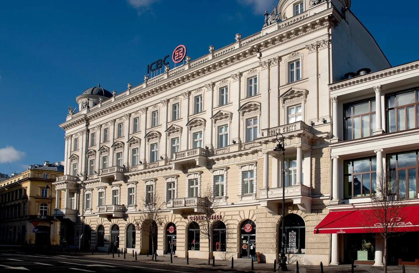 White historic building with decorative architectural details and large windows, with a red awning and a coffee shop on the ground floor, under a clear blue sky.
