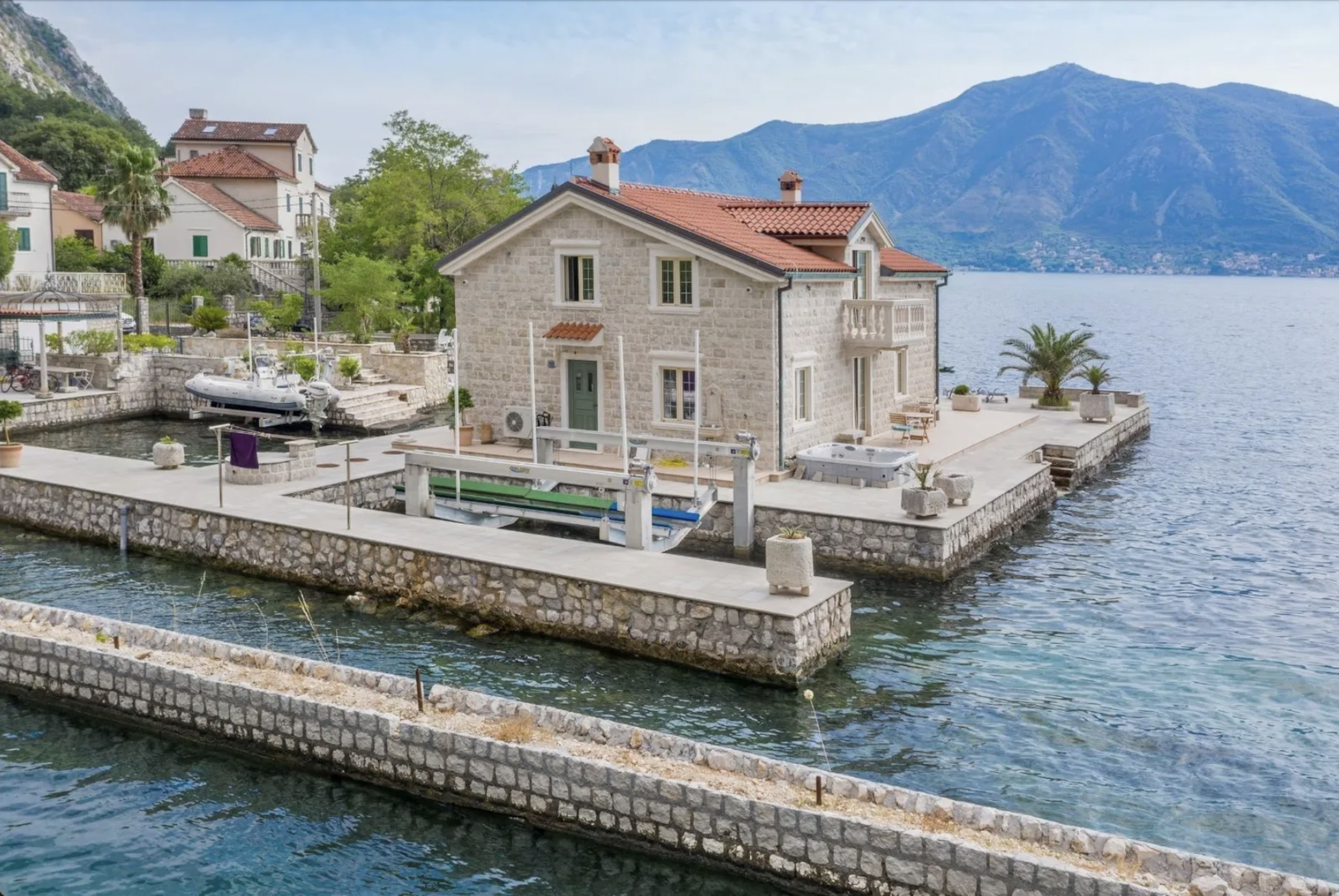 Stone house on a pier by a lake with boats, mountains in the background, and a clear sky.