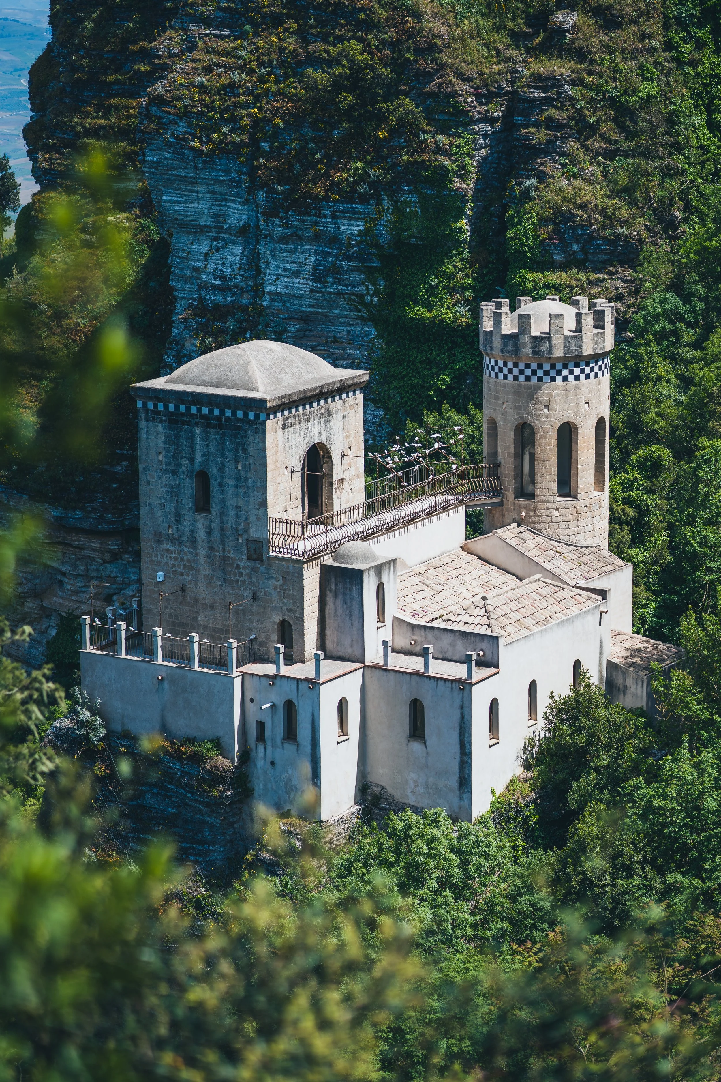 Castle-like building on a hillside with a tower, surrounded by lush green trees and rocky cliffs in the background.