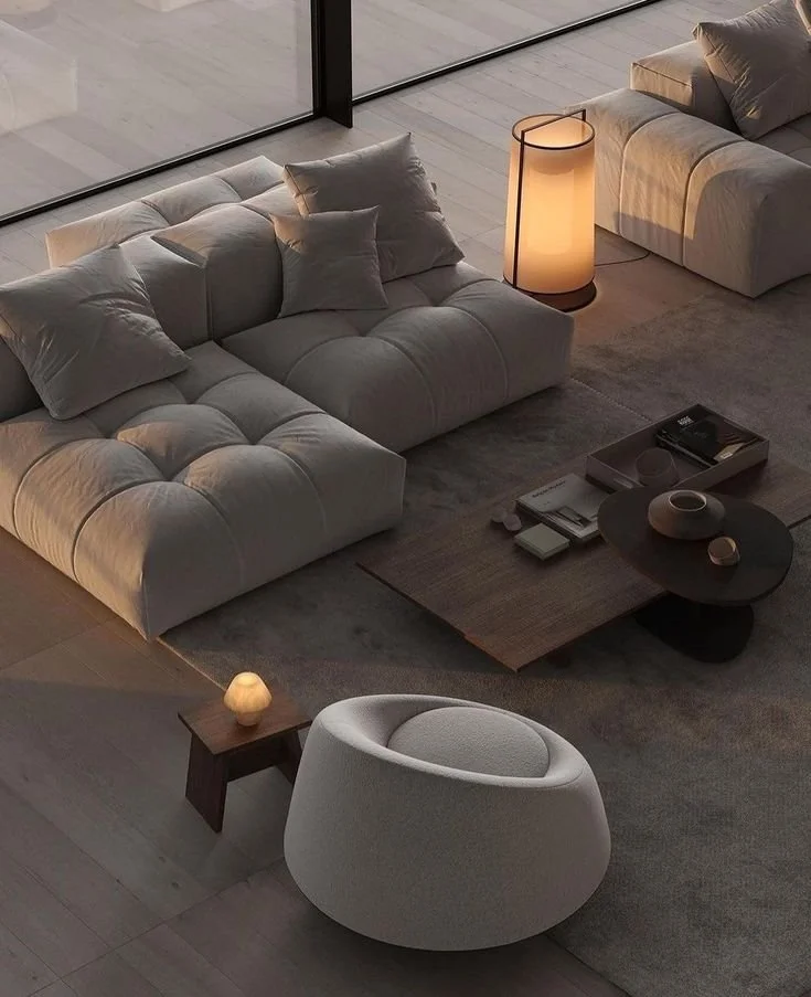 Modern living room with light-colored sofas, a round white armchair, a side table with a small lamp, and a dark coffee table with decor items, illuminated by warm lighting near floor-to-ceiling windows.