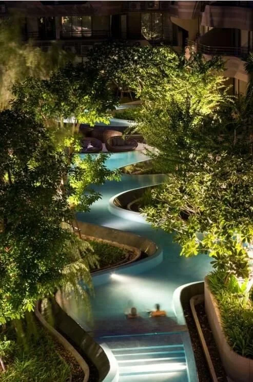 Night view of a winding outdoor pool surrounded by lush trees and greenery, with some people swimming in the pool.