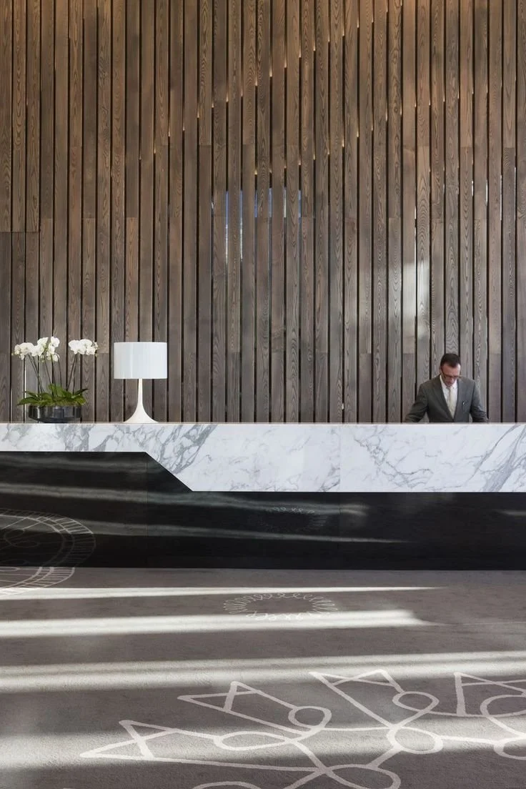 Modern hotel lobby reception area with a marble front desk, a table lamp, orchid flowers, and a man in a suit standing behind the desk. Wooden panel wall in the background with sunlight casting shadows on the floor.